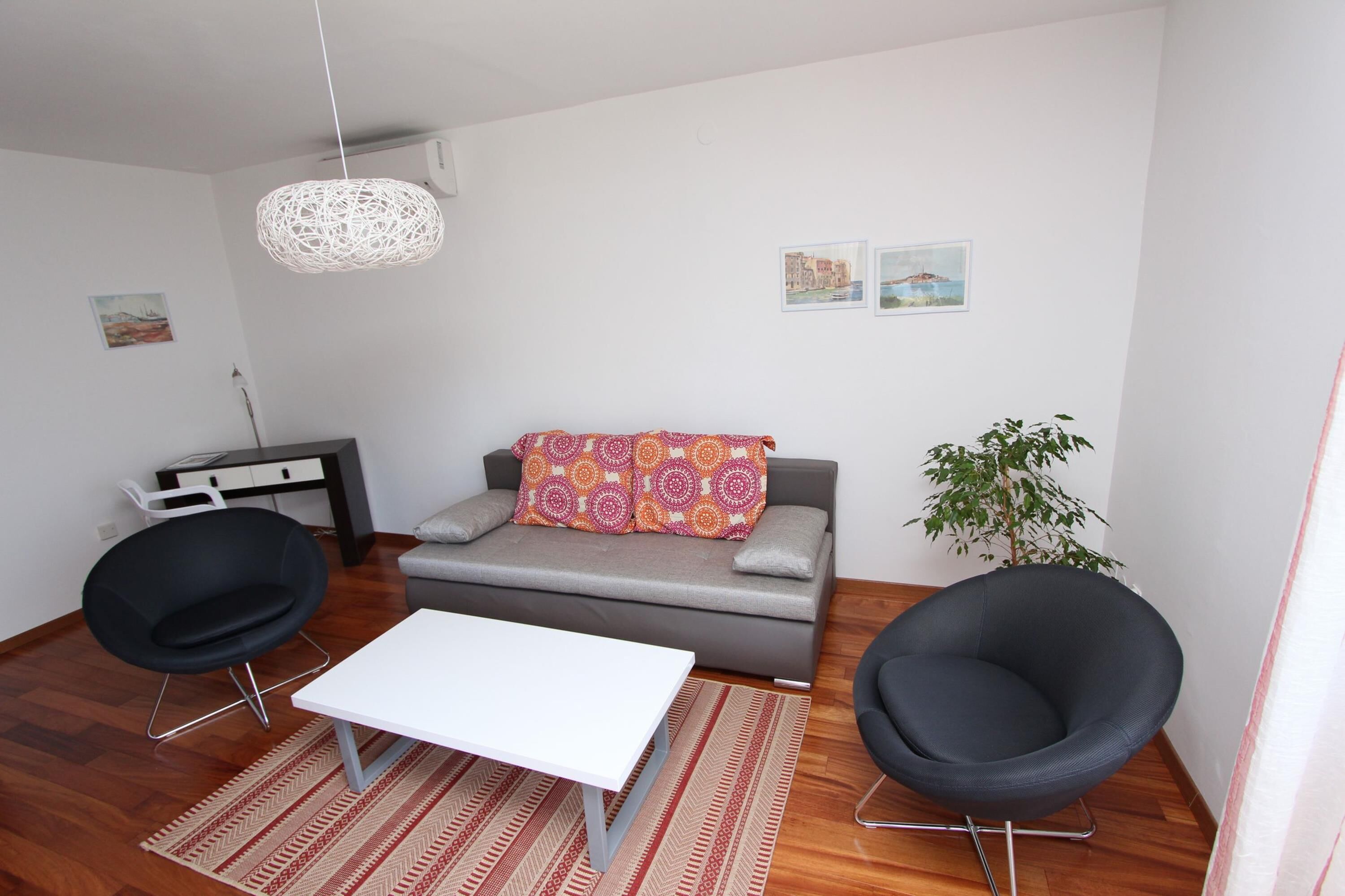 A modern living room with a grey sofa, two black chairs, a white table, a patterned rug, and a hanging light fixture.