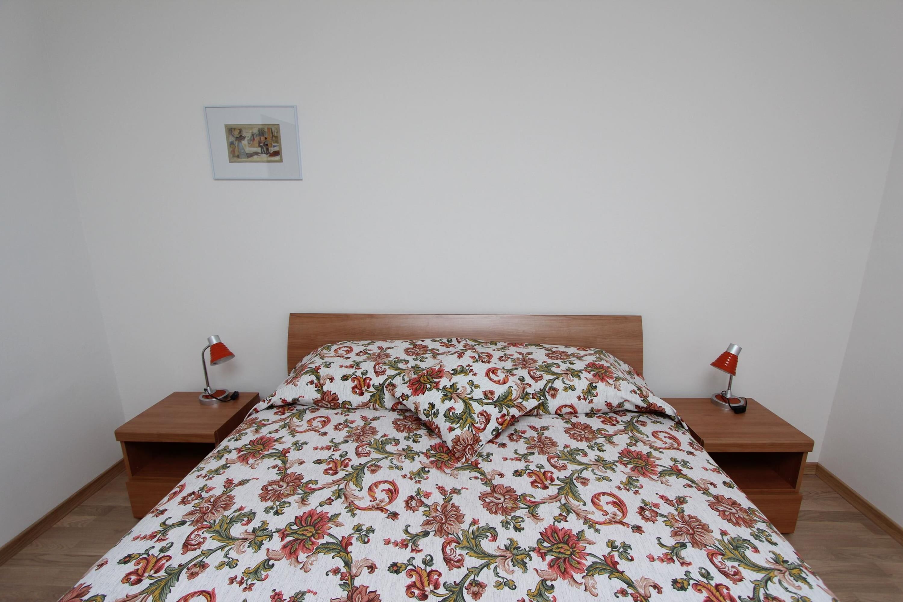 Ein Schlafzimmer mit floralem Bettbezug, zwei Nachttischen mit Lampen und einem gerahmten Bild an der Wand.