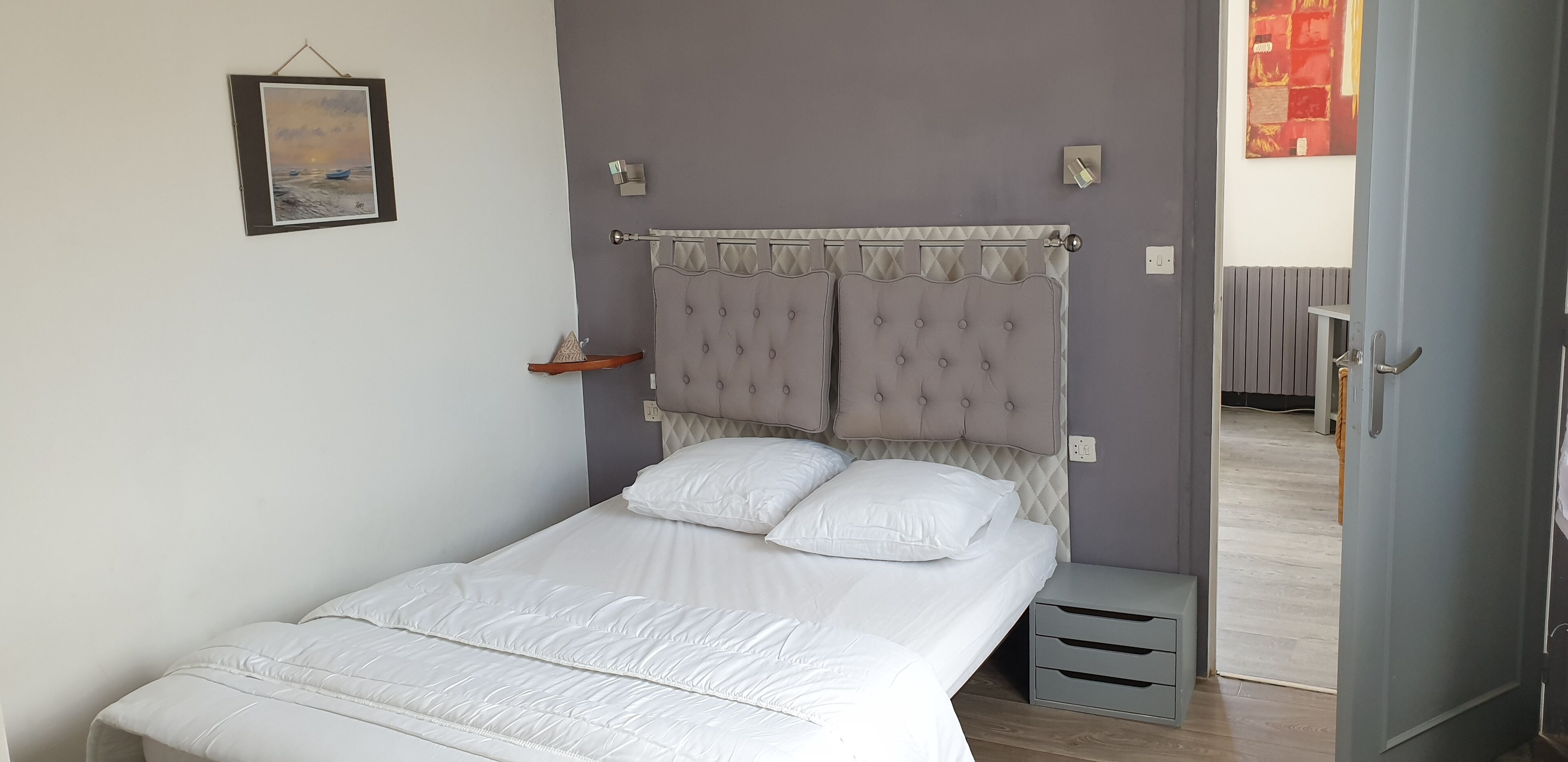 Een net opgemaakt bed met wit beddengoed in een kamer met een grijze muur, een lijst met een afbeelding en een deur naar een andere kamer.