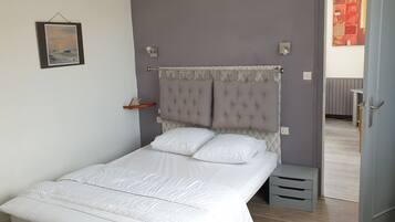 Een net opgemaakt bed met wit beddengoed in een kamer met een grijze muur, een lijst met een afbeelding en een deur naar een andere kamer.