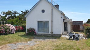 A white single-story house with a large window and a door, surrounded by a garden with pink flowers and a gravel path. - Pretty villa in Trévignon, 2 steps from the beach and port (Trégunc)