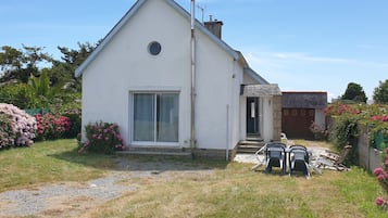 Een wit huis met één verdieping, met een groot raam en een deur, omgeven door een tuin met roze bloemen en een grindpad.