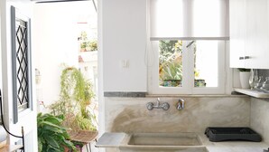 Une cuisine avec des meubles blancs, un plan de travail en marbre, un évier et une fenêtre donnant sur un balcon agrémenté de plantes.