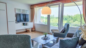 Ein modernes Wohnzimmer mit einem Flachbildfernseher, einer Couch, einem Couchtisch und einem großen Fenster mit Blick auf Grünflächen.