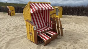 Ein Liegestuhl mit gestreiftem Sonnenschirm, beschriftet mit K 277, am sandigen Strand.