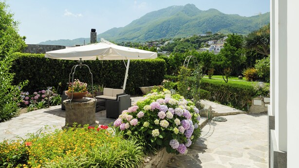 A patio with a white umbrella, outdoor seating, and a flower bed. - Historic Charm with Modern Comfort near the Sea (Lacco Ameno)