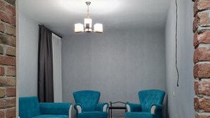 Een kamer met een bakstenen muur, een houten vloer, een grijze plafond en een kroonluchter. Er staan twee blauwe fauteuils en een bank.