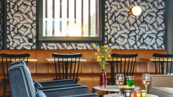 A restaurant interior with a patterned wall, a window with vertical blinds, and a table with a vase of flowers, wine glasses, and condiments.