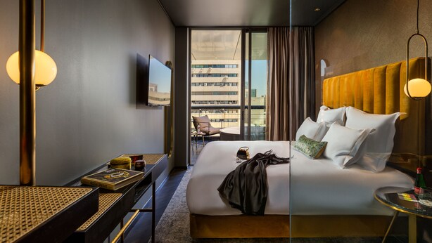A modern hotel room with a large bed, a TV, a desk with a lamp, and a view of the city through large windows. - Brown Machne Yehuda (Jerusalem)