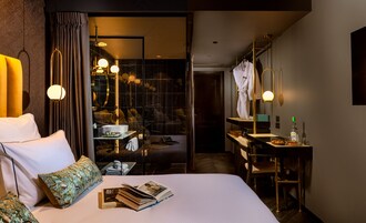 Habitación de hotel con cama, escritorio, espejo y baño visible a través de un panel de vidrio.