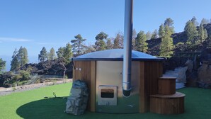 Eine moderne Sauna im Freien mit Schornstein, umgeben von einer Holzkonstruktion und einer Rasenfläche.