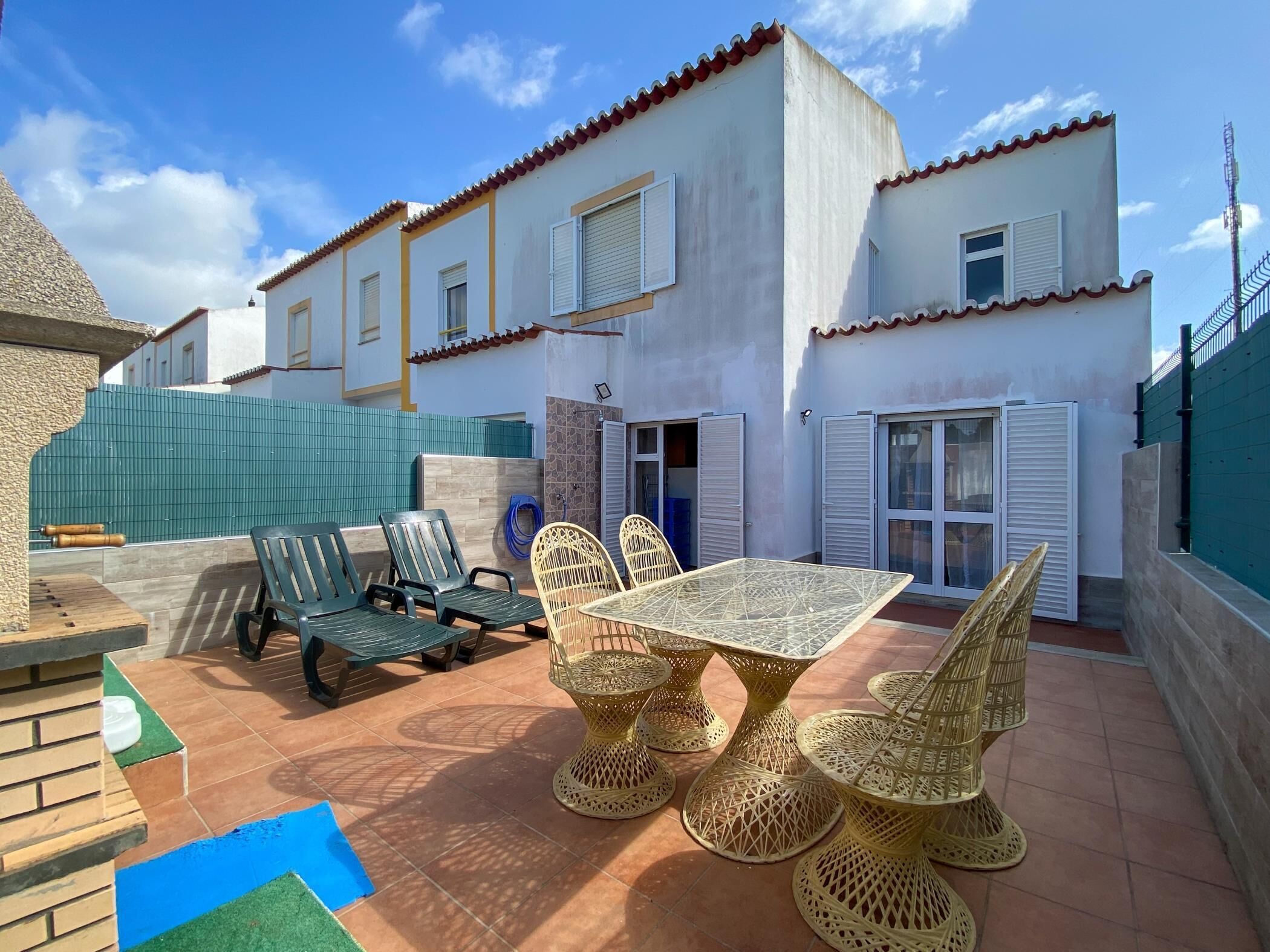 Une maison à deux étages avec un patio comprenant une table et des chaises, une cl ôture verte et un tapis bleu.