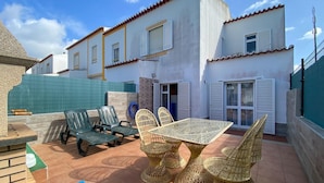 Una casa de dos pisos con un patio que incluye una mesa y sillas, una cerca verde y un tapete azul.