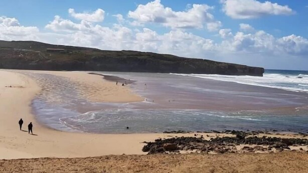 Una playa con un promontorio rocoso al frente y una playa arenosa con olas suaves.
