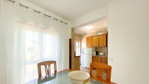 Un comedor con una mesa de vidrio, sillas y un cuenco. Se observa una cocina al fondo con armarios de madera y un refrigerador.
