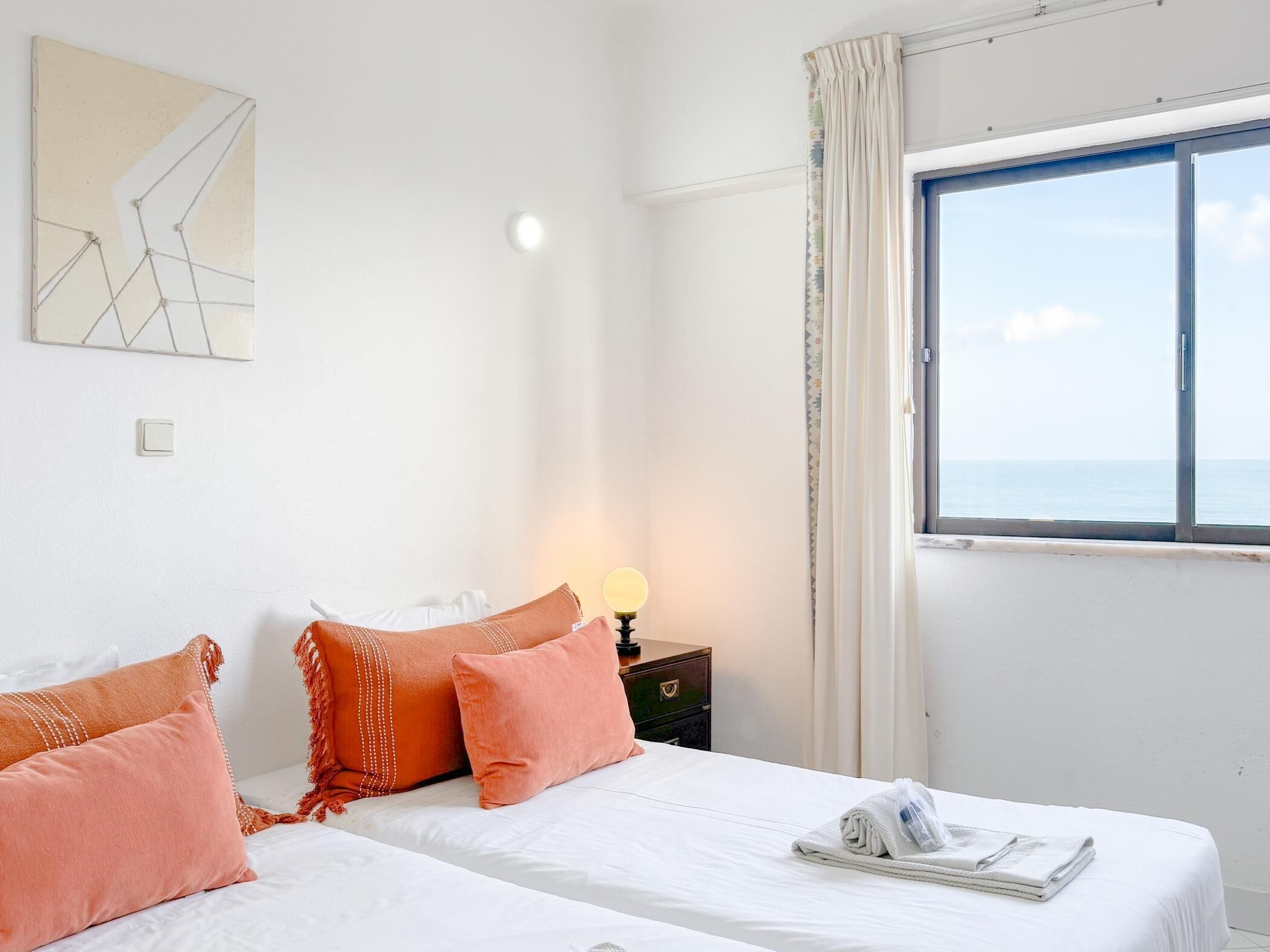 Ett hotellrum med en säng, kuddar, ett nattduksbord, en lampa, ett fönster med utsikt och ett konstverk på väggen.
