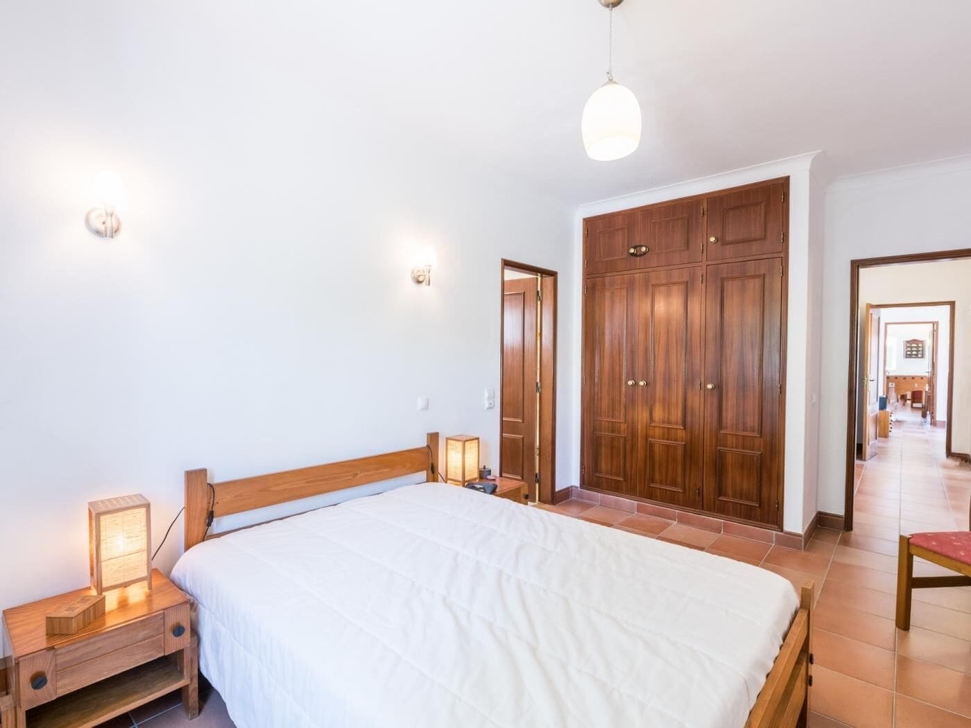Un dormitori amb un llit de fusta, taules de nit amb làmpades, un armari i un passadís que condueix a una altra habitació.