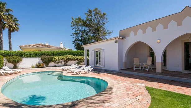 Ein Pool mit gewölbter Kante, umgeben von einer Ziegelterrasse und einer Rasenfläche. Liegestühle und ein kleines Gebäude mit Bögen sind ebenfalls vorhanden.