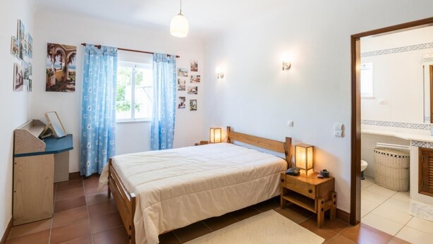 Quarto com cama, abajures, mesa de cabeceira de madeira, escrivaninha, janela com cortinas e luminárias na parede.