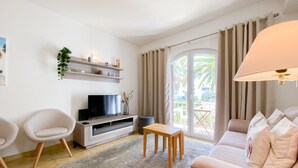 Een moderne woonkamer met een flatscreen-televisie, een bank, twee fauteuils, een klein tafeltje en een balkon met gordijnen.