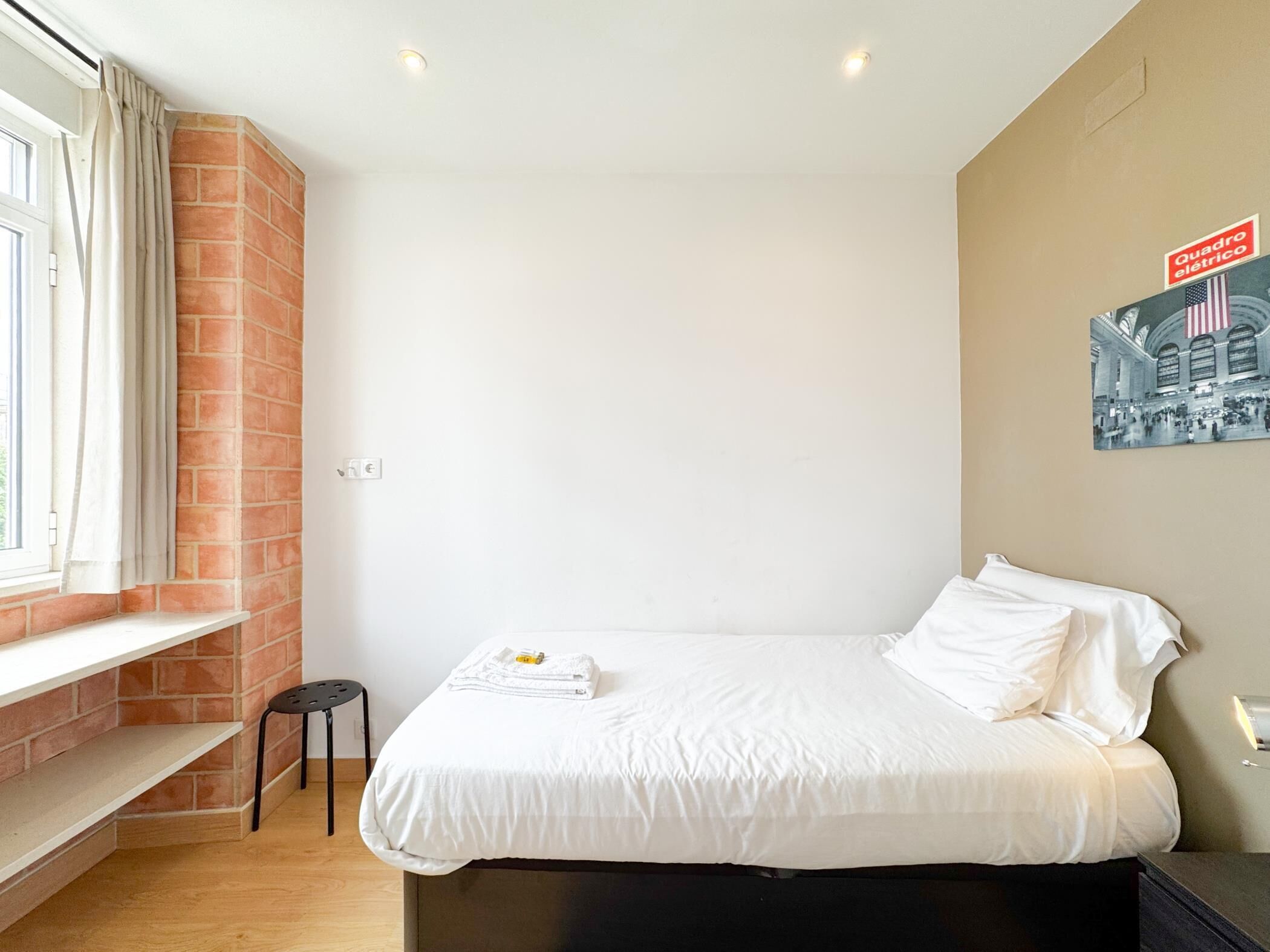A hotel room with a bed, a window with curtains, a brick wall, a small stool, and a poster on the wall.