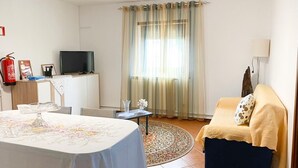 A well-lit room with a bed, a sofa, a television, a fire extinguisher, and a window with curtains.