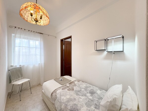 A bedroom with a bed, a chair, a window with curtains, a door, and a wall-mounted light fixture.