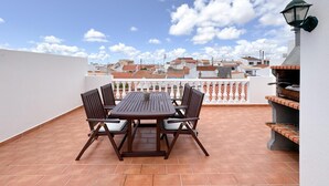 A rooftop terrace with a wooden table and chairs, a grill, and a view of the cityscape.