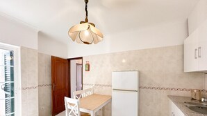 Une cuisine avec un réfrigérateur blanc, une table à manger en bois avec des chaises blanches, un luminaire suspendu et une fenêtre avec des stores.