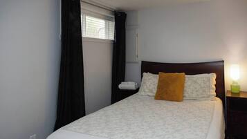 Een enkel bed met hoofdeinde, een nachtkastje met lamp, een raam met gordijnen en een wandgemonteerde airconditioner.