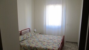 A small, single-bed room with a floral bedspread, a nightstand, and a window with sheer curtains. - SEASIDE APARTMENT (Alcamo Marina)