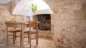 Une pièce rustique aux murs de pierre, avec une table et des chaises en bois, un four en briques blanches en forme d’arche et une plante suspendue.