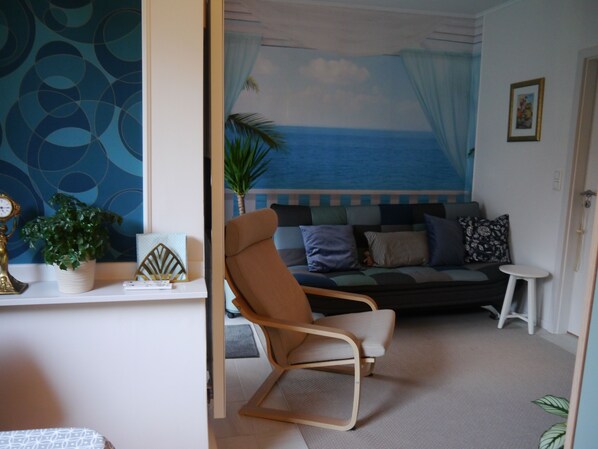 A room with a beach mural, a sofa, a chair, a small table, and a potted plant. - Blaue Welle - Gemütlichkeit mit Stil (Varel)
