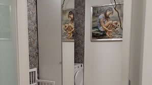 A compact bathroom with a white sink, a mirror, a small white bench, a washing machine, and two framed artworks on the wall. - Blaue Welle - Gemütlichkeit mit Stil (Varel)