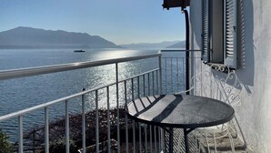 A balcony with a metal railing overlooking a body of water, a small round table, and a building with shutters. - Appartamento Ca Bianca  (Cannobio)