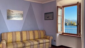 A room with a sofa, a window with a view of a body of water and buildings, and two framed pictures on the wall. - Appartamento Ca Bianca  (Cannobio)