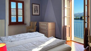 A bedroom with a bed, a chair, a dresser, and a window with a view of a body of water. - Appartamento Ca Bianca  (Cannobio)
