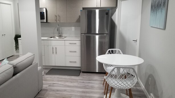 Cocina compacta con refrigerador de acero inoxidable, armarios blancos y una pequeña mesa de comedor con sillas blancas.