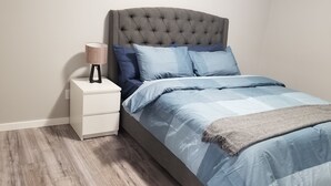 Un dormitorio con cabecera capitonada, una mesita de noche con lámpara, y una cama con ropa de cama azul y una manta gris.