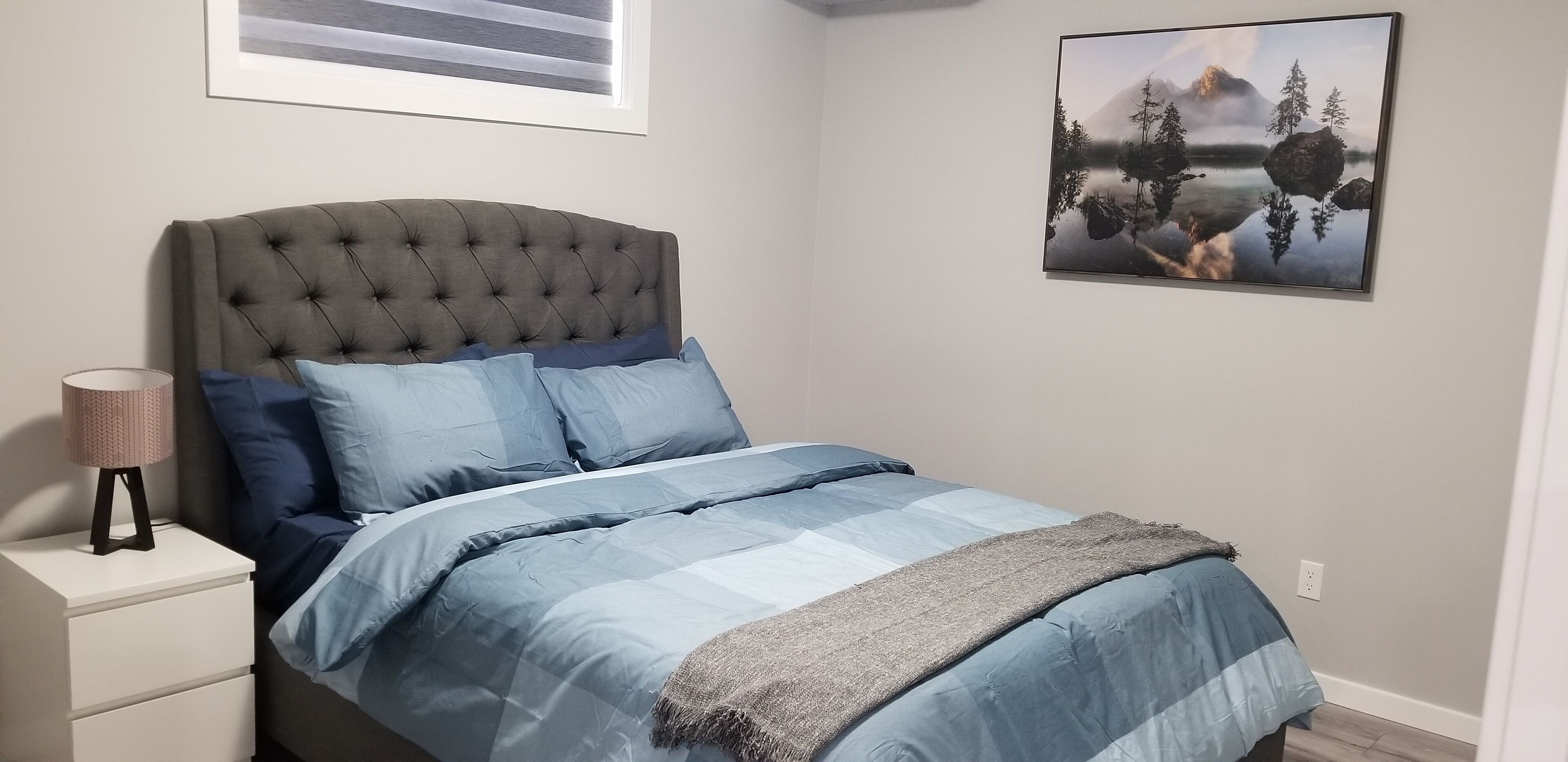 A bedroom with a tufted headboard, a bed with blue bedding, a bedside lamp, and a framed landscape painting on the wall.