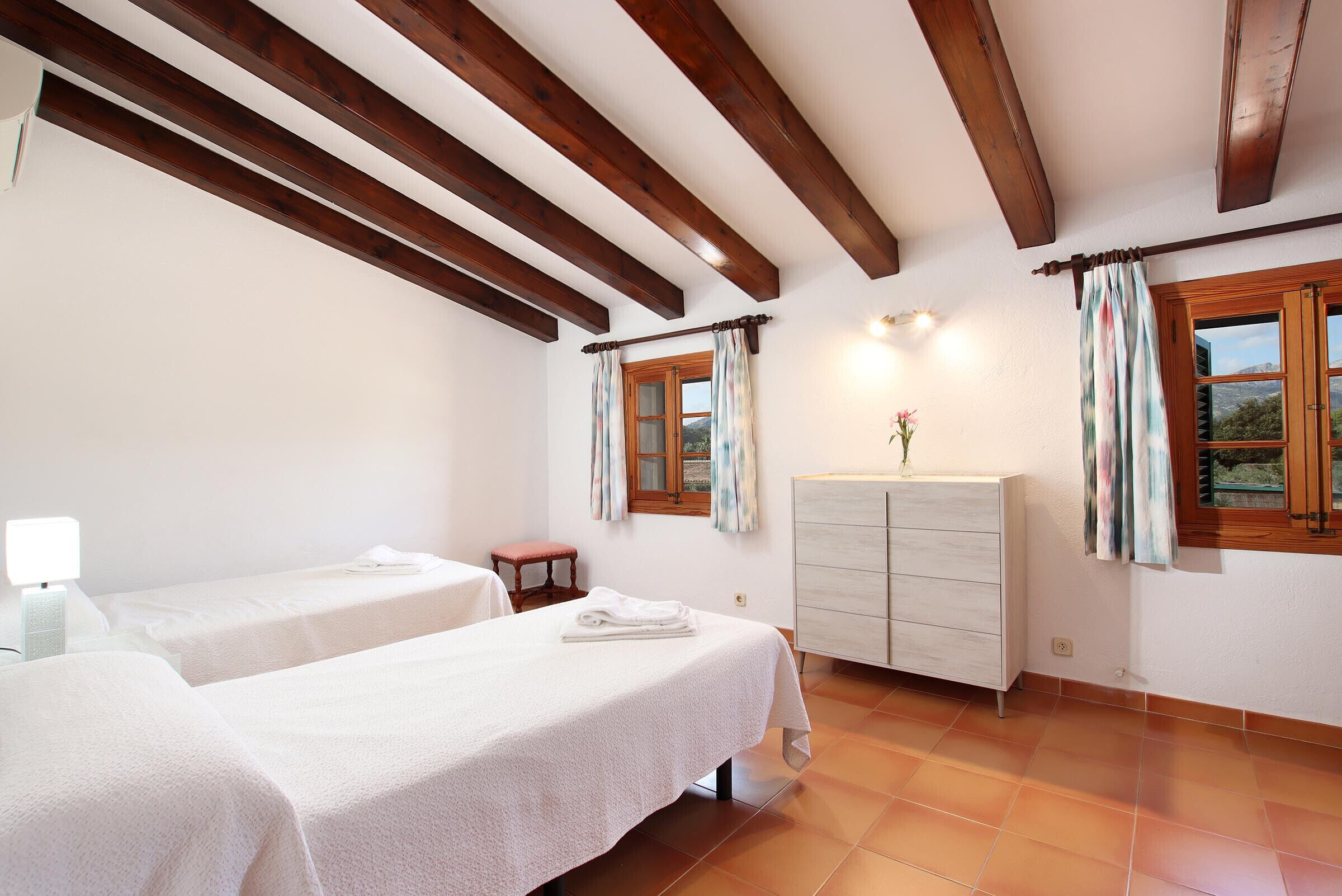 A room with two beds, wooden beams on the ceiling, a dresser, and a window with curtains.