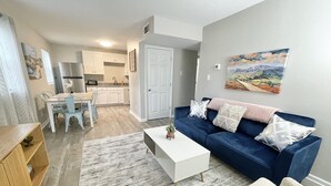 Een moderne woonkamer met een blauwe bank, een witte salontafel en een grijs vloerkleed. Er is een eethoek met een houten tafel en stoelen. De keuken is zichtbaar op de achtergrond, met witte kasten en een grijs aanrecht.