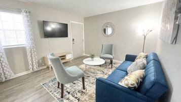 Un salon avec un canapé bleu, une table basse ronde, un fauteuil gris et un téléviseur fixé au mur.