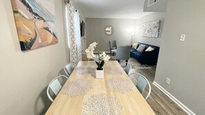 Un coin repas avec une longue table en bois, des chaises blanches et des sets de table décoratifs. On aperçoit un canapé bleu et un téléviseur au fond.