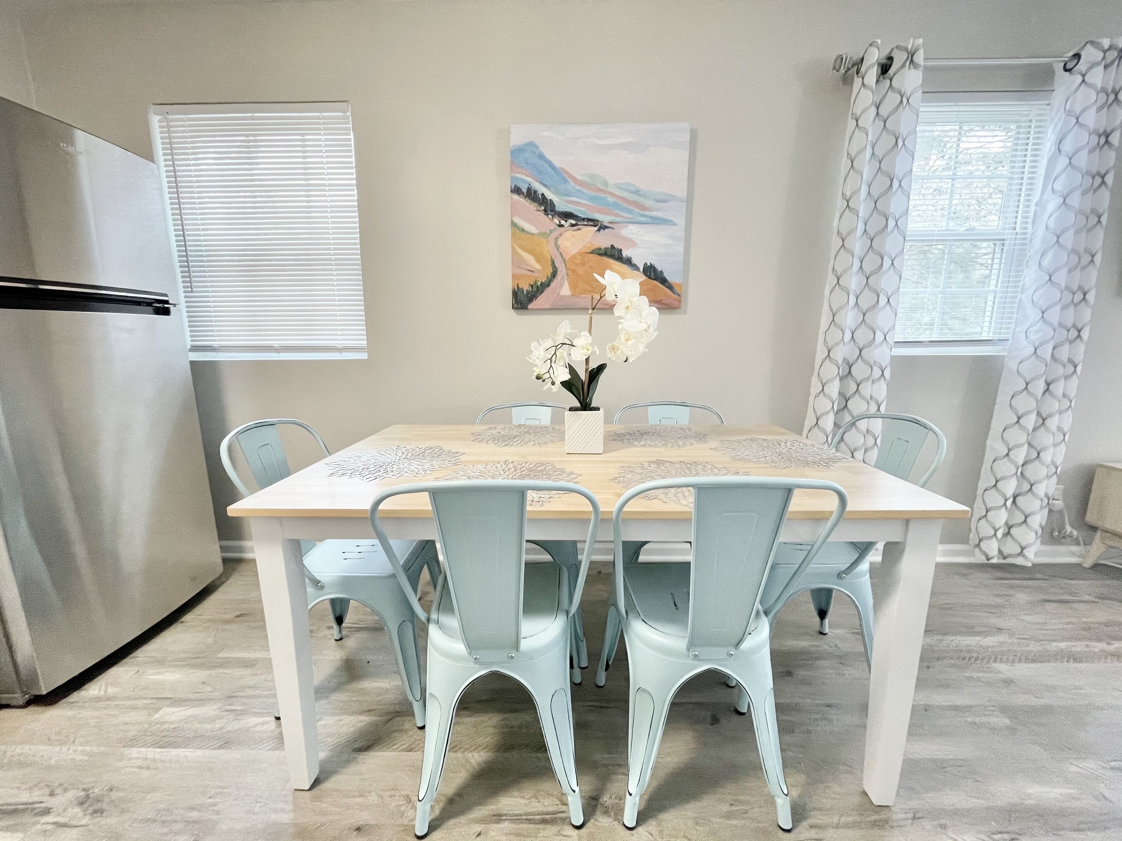 Un coin repas avec une table en bois, des chaises bleu clair, un vase blanc orné de fleurs et un tableau accroché au mur.