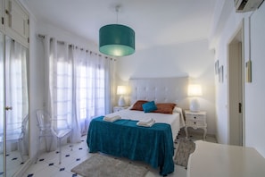 Une chambre à coucher avec un lit, deux tables de chevet, une chaise transparente, une lampe de couleur turquoise et un plafond blanc.