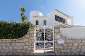 Un bâtiment blanc avec un mur en pierre, un portail en métal et un palmier.