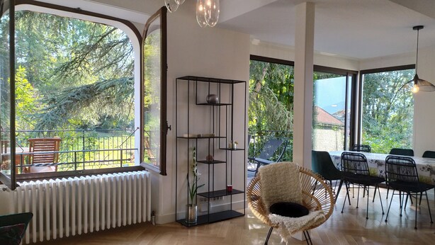 Amplia sala de estar con ventanas grandes, un radiador, un estante negro y una silla de mimbre.