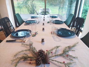 A dining table set for six with dark blue plates, clear glasses, and a centerpiece of pine branches and pinecones.