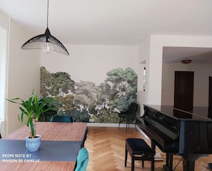 A modern living room with a piano, a dining table with chairs, and a large wall mural.
